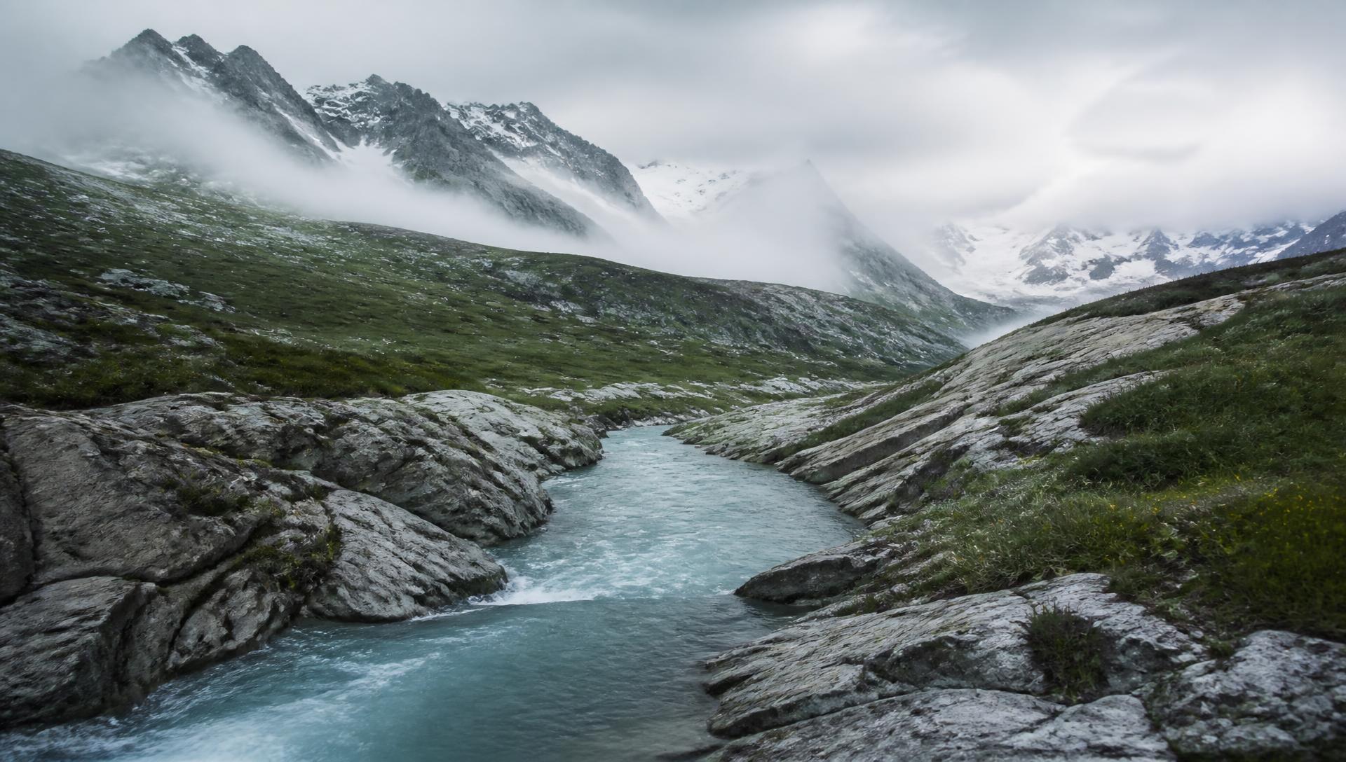A glacial alpine river running between rock and meadow beneath misted Swiss peaks